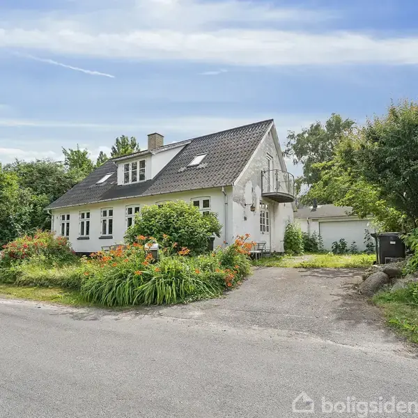 Hvidt hus med mørkt tag står i stille villakvarter. Blomsterbuske omgiver indkørslen, mens en garage ses i baggrunden under en blå himmel.