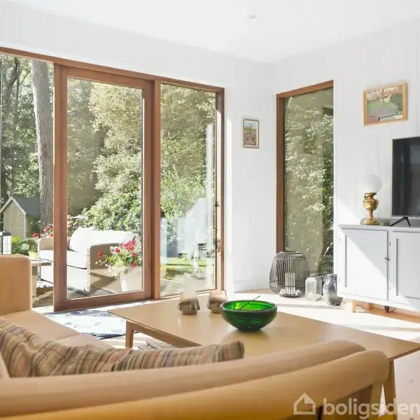 Stue med beige sofa, der vender mod et træbord med grøn skål. Store vinduer viser en frodig have. Til højre står et TV på en grå skænk.