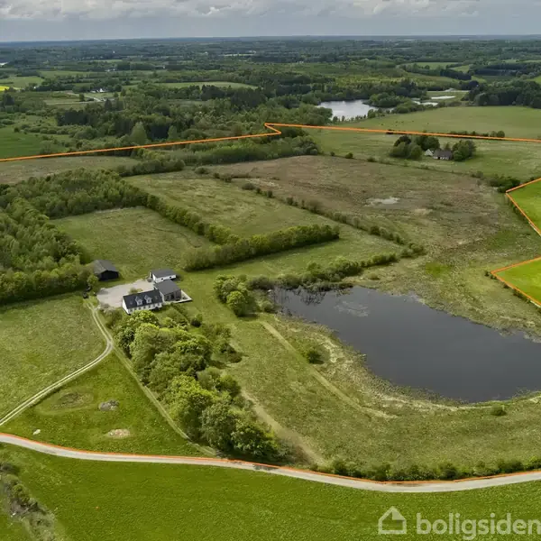 Et hus ligger midt i en stor, afgrænset landlig ejendom med en sø og åbne marker. Omkringliggende skovområder og små bakker breder sig under en overskyet himmel.