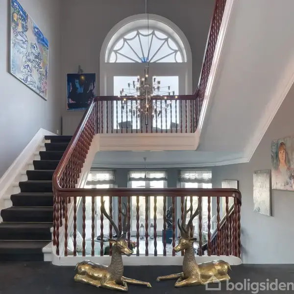 To guldrensdyr sidder på tæppet i en elegant hall med en bred trappe, mørkt trægelænder og kunstmalerier på væggene, oplyst af en stor lysekrone og buet vindue.