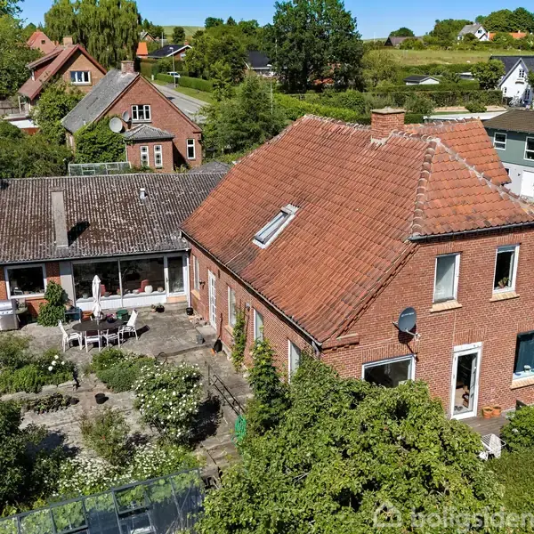 Rødt murstenshus med rødt tegltag omgivet af en frodig have. Der er flere mindre huse i nabolaget på en solrig dag med grønne træer og buske omkring.