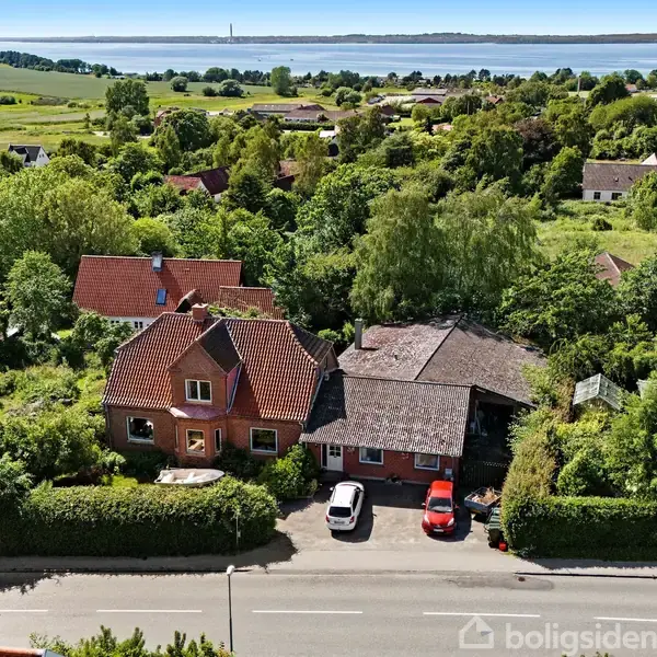 Et rødt hus med to biler i indkørslen er omgivet af frodige træer og buske i et landligt område nær en vej. I baggrunden ses en fjord og marker.