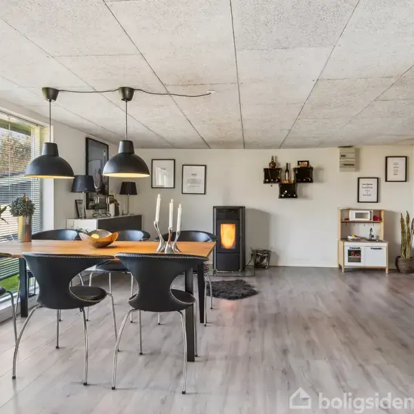Spisebord med sorte stole står i stue med store vinduer og trælaminatgulv. En pejs er tændt, og vægudstillinger inkluderer billeder. Lysekrone hænger over bordet.