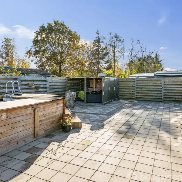 Træbassin står stationært på flisebelagt terrasse omgivet af træhegn. I baggrunden ses et lille skur og træer, mens der er nogle havemøbler til højre.