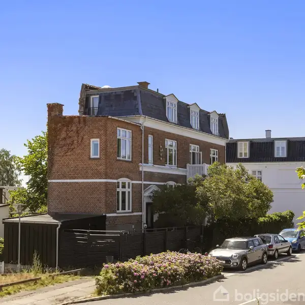 Bygning med røde mursten og sorte tage står stille. Flere biler er parkerede langs vejen i et beboelsesområde med træer og buske i nærheden.