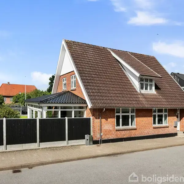 Et rødt murstenshus med hvidt bindingsværk og skråt tag står på en gade med flere huse. Huset har store vinduer og en indhegnet terrasse i forhaven.