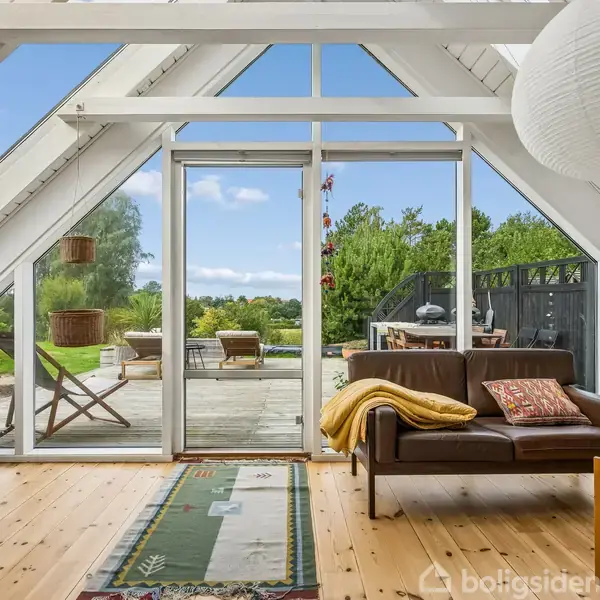 Lædersofa med puder står i en stue med højt til loftet og store glaspartier, der fører ud til en terrasse med havemøbler og grøn udsigt.