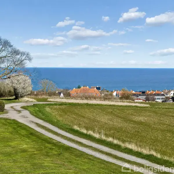 En snoet grusvej løber gennem en grøn mark, med træer i baggrunden. I horisonten ses røde hustage og en blå havudsigt under en klar, skyet himmel.