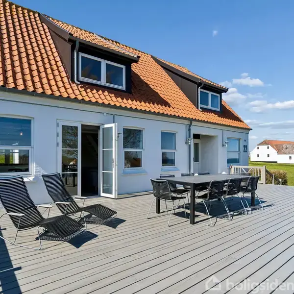 Hus med rødt tegltag og to kviste. To liggestole og et bord med stole står på en træterrasse. Omkringliggende græsareal og andet hus i baggrunden under blå himmel.