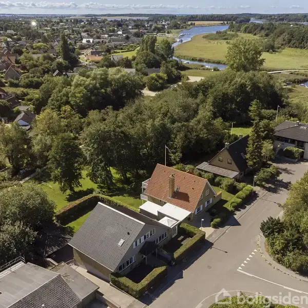 Rødt tegltagshus omgivet af grønne træer i et villakvarter nær en flod. Flere huse ses, og landskabet strækker sig mod horisonten under en skyfri himmel.