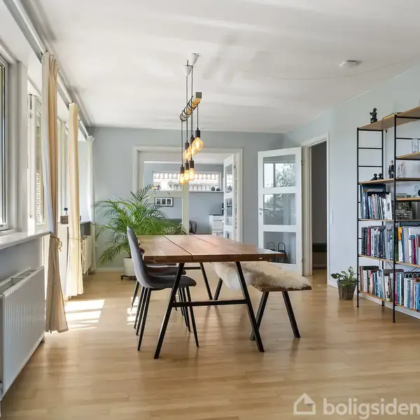 Et spisebord i træ med sorte stole omkring står i en lys stue med trægulv. En reol fyldt med bøger står til højre, og et vindue med gardiner lader dagslys strømme ind.