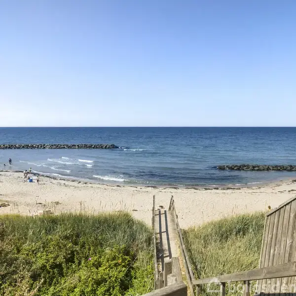 Trætrappe fører ned til sandstrand med personer i aktivitet ved havet; omgivet af grønne klitter og blåt hav under klar himmel.