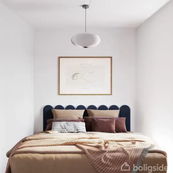 En seng står centralt med mange puder og en dyne. Over sengen hænger et kunstbillede i ramme. Værelset har hvide vægge og en loftslampe.
