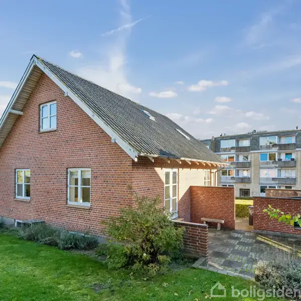 Et rødt murstenshus står stille i en velplejet have med græs og buske. Bagved ses en moderne lejlighedsbygning. Himlen er klar og blå.