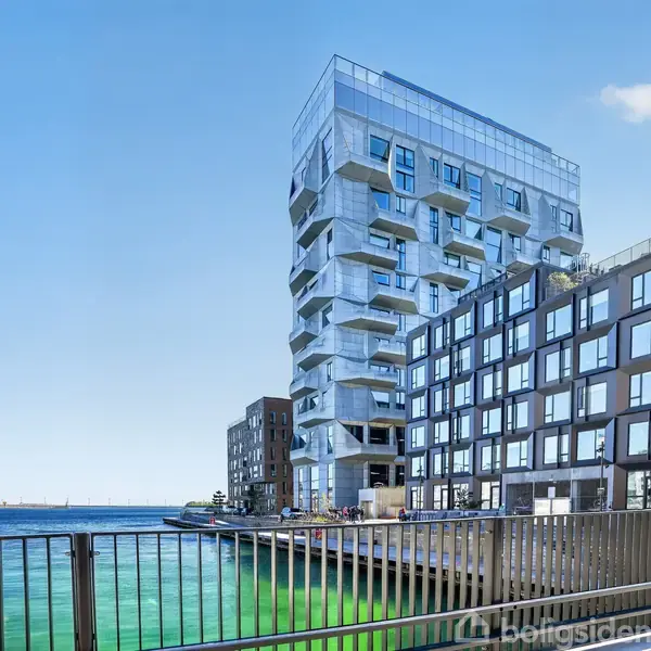 Et moderne, kantet lejlighedskompleks med store vinduer står opført ved en kanal med klart grønt vand, omgivet af andre moderne bygninger under en blå himmel.