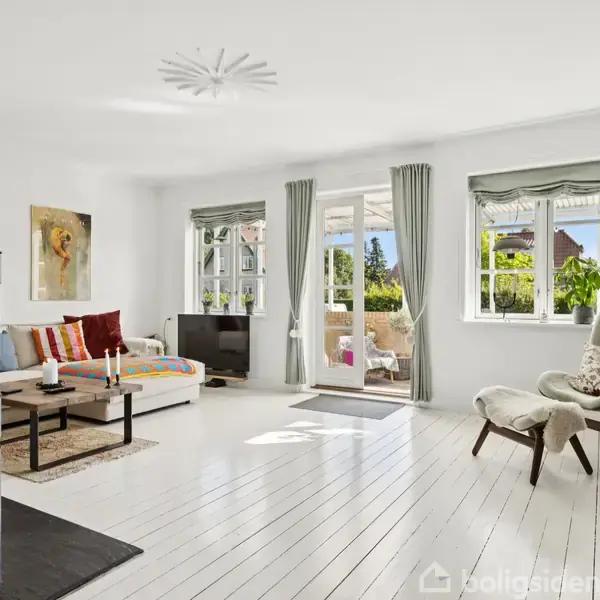 Stue med pejs i aktion, lyst trægulv, beige sofa, og lænestol. Vinduer og glasdør viser udsigt til grønt haveområde. Reol med planter og billeder til højre.