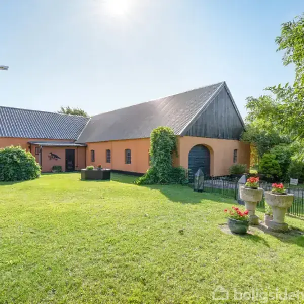 Et orange hus med sort tag står på en grøn græsplæne. Tre blomsterpotter med blomster ses i forgrunden. Sollys skinner, og der er træer i baggrunden.