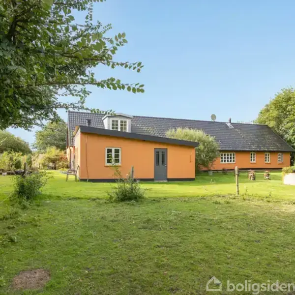 Et gult hus med sort tag står i en grøn have omgivet af træer og buske på en solrig dag.