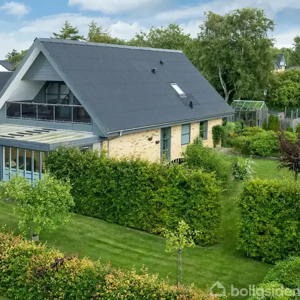 Et moderne hus med skråt tag står omgivet af frodig vegetation. Glaspaneler ses på terrassen, mens grønne buske og træer omkranser ejendommen i en rolig forstadsmiljø.