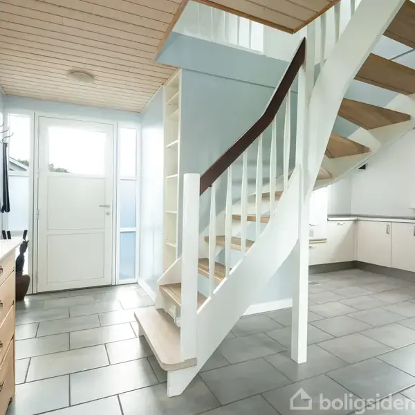 En hvid trappe drejer opad mod overetagen i et lyst rum med beige flisegulv. Omkring trappen er der køkkenskabe, et vindue og en kommode med planter.