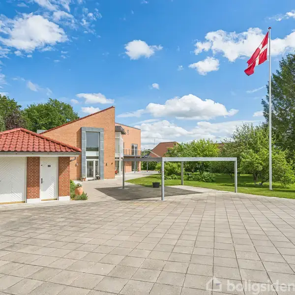 En moderne murstensvilla med store vinduer står ved siden af en garage. En dansk flag vifter i vinden, omgivet af en velplejet have og flisebelægning.