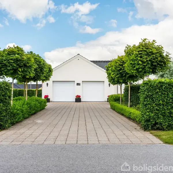 Hvidt hus med to garageporte står for enden af en brostensbelagt indkørsel. Store grønne træer og velplejede hække flankerer indkørslen under en delvist skyet himmel.