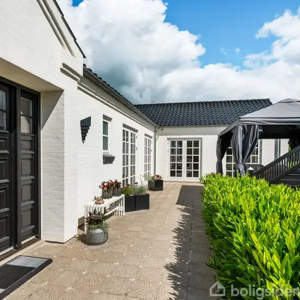 Et hvidt hus med sorte vinduer og døre står i solen. En terrasse med grønne buske og en pavillon skaber en hyggelig udendørs atmosfære under en blå himmel med skyer.