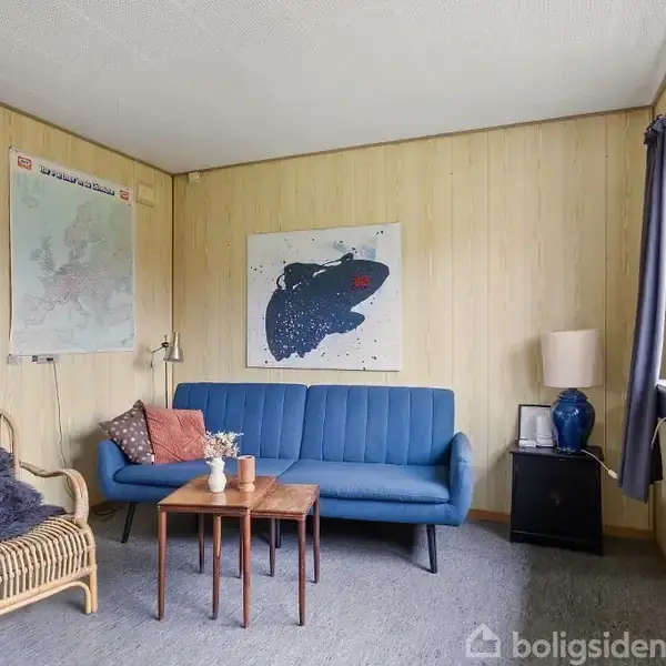 En blå sofa står i en stue med trægulv og lyse trævægge. På væggen hænger et kort og et maleri. En bambuslænestol og et lille bord står ved siden af.