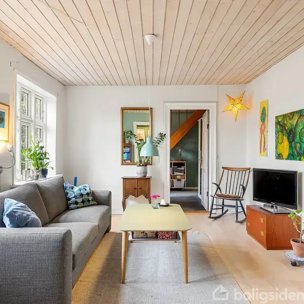 Stue med grå sofa til venstre og tv på skænk til højre. Et lysegult sofabord står foran sofaen. Rummet er lyst med træloft og dekorerede vægge.