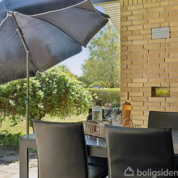 En sort parasol skygger et bord dækket med glas og en kande udenfor. Konteksten er en terrasse ved siden af en gul murstensvæg, med udsigt til en have.