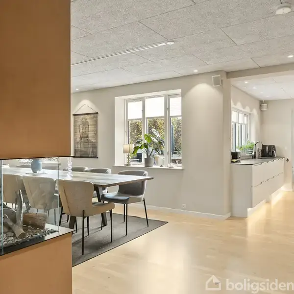 En moderne spisestue med et bord og stole placeret ved siden af store vinduer, der slipper naturligt lys ind. I baggrunden ses et hvidt køkken med lange bordplader.