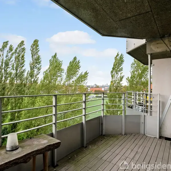 Balkon med træplanker og metalrækværk; en bænk står til venstre. Træer blæser i vinden i baggrunden under en blå himmel med skyer.