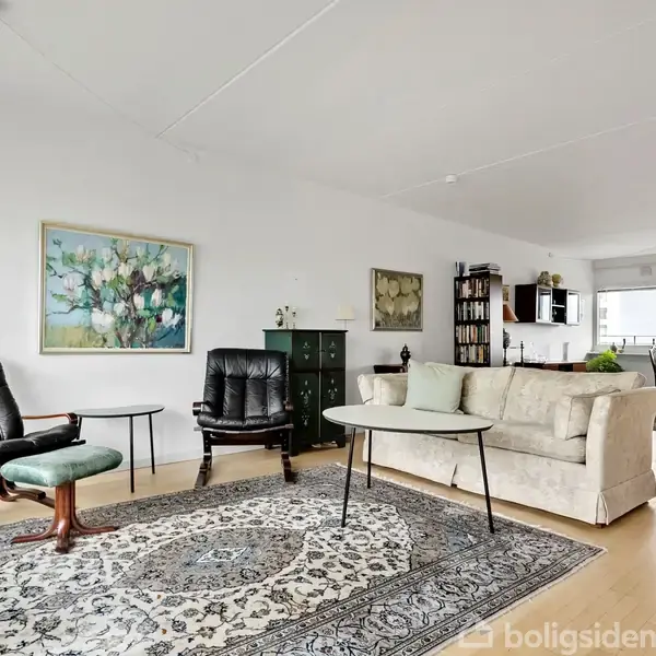 Stue med to lænestole og et sofabord på et orientalsk tæppe; sofa ved siden af. Billeder hænger på væggen, og spisebord ses i baggrunden i et lyst rum.