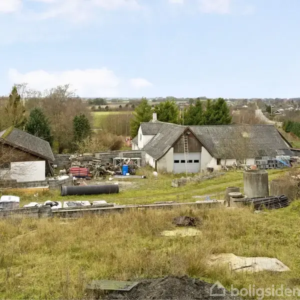 Landbrugsejendom med flere bygninger står stille omgivet af åbne marker og træer. Bygningerne synes ubrugte, og omgivelserne er prægede af græs og opbevarede materialer.
