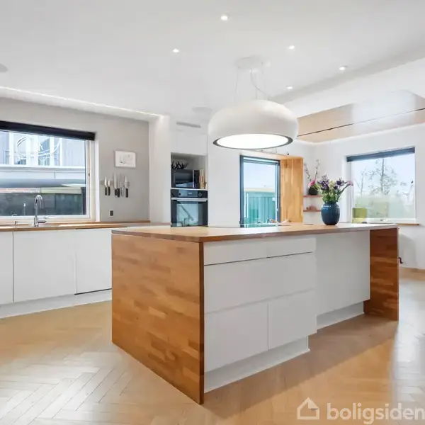 Et moderne køkken med hvidt interiør og træbordplader. En stor ø med indbygget lys hænger over. Rummet har flere vinduer der lukker naturligt lys ind.