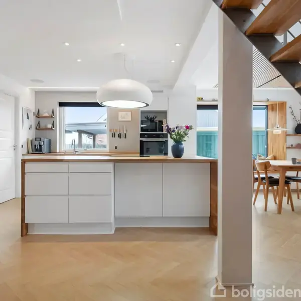 Hvidt køkken med ø, vase og ovn set i baggrunden, stilfuldt integreret med spiseplads til højre, med trappeopgang. Lyse omgivelser med trædetaljer og moderne møblering.