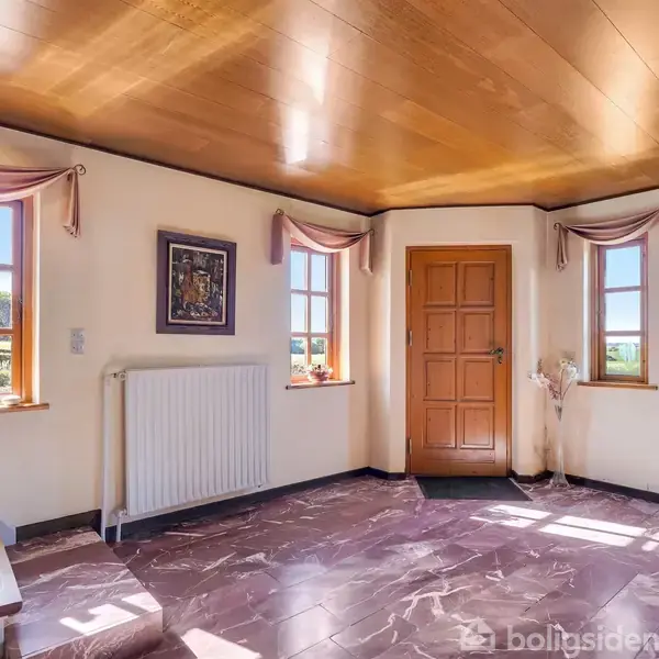 Entré med trægulv og træloft, tre vinduer med lyserøde gardiner, og en trædør. Der er en radiator under et vægmaleri, en kommode til højre og en vindeltrappe til venstre.