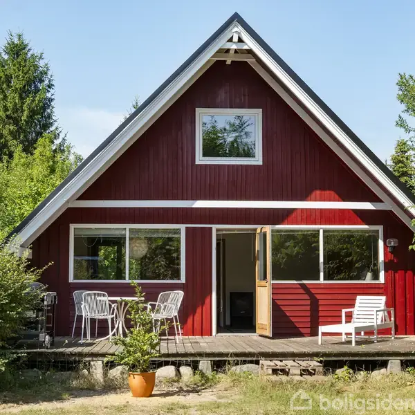 Rødt træhus med spidst tag står stille på en grøn havegrund. Åben dør til terrasse med havemøbler. Omgivet af træer og buske i solrigt vejr.