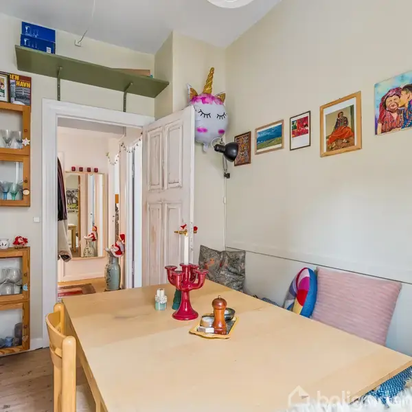Et spisebord med stearinlys og dekorationer står i et lille rum med væghylder, farverige puder og kunstværker på væggen. En dør fører til et tilstødende rum.
