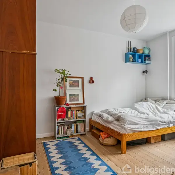 Seng med ureddet sengetøj står mod en hvid væg i et lyst soveværelse. Omkring er et trægulv, blå tæppe, en reol med bøger og planter, samt et vindue.