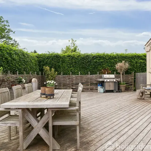 Langt træbord med stole står på en træterrasse. Grill og planter omringer dem. Høje hække omkranser terrassen, mens en blå himmel ses i baggrunden.