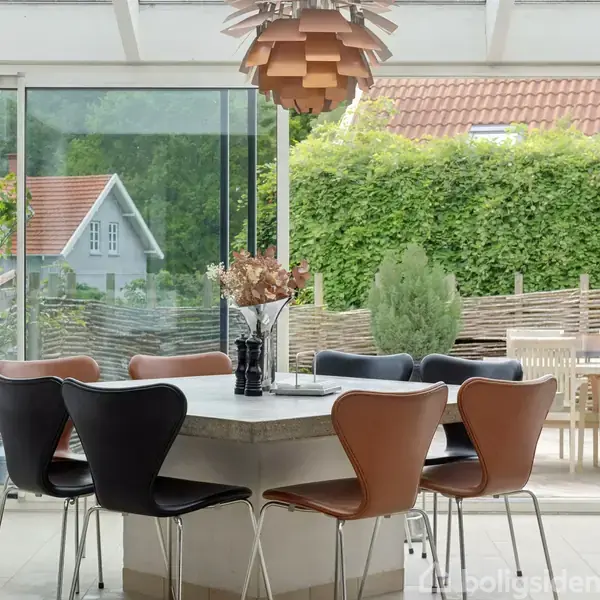 Et moderne spisebord med otte stole i sort og brun står i et lyst rum med glasvægge. En dekorativ lampe hænger over bordet. Udsigt til en have med træhegn.