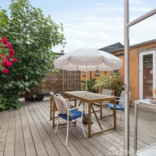En træterrasse har et bord med stole under en hvid parasol. Omkranset af røde blomster og grønne buske ved et gult, murstenshus.