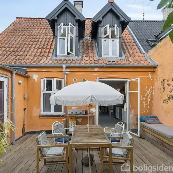 Et bord med parasol og stole står i midten af en træterrasse foran et charmerende gult hus med skråtag og åbne døre, omgivet af planter og røde blomster.