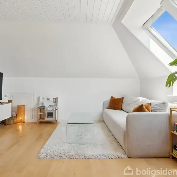 TV'et står på et hvidt møbel, sofaen er placeret ved siden af et glasbord, og rummet har skråvinduer og træplante i en moderne stueindretning med lyse vægge.