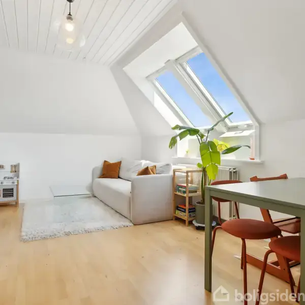 En lille spisestue med to træstole står ved et bord ved en skråvæg. En stor plante og en buket blomster i en vase skaber en hyggelig atmosfære.