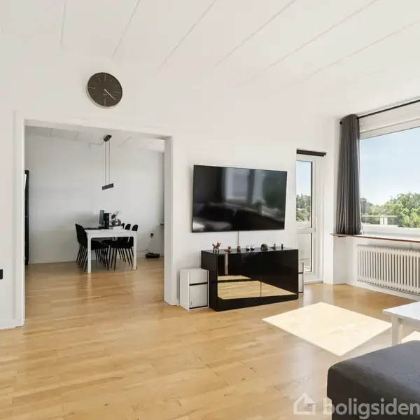 Stue med trægulv og vægmonteret fladskærms-tv. Rundt vægur hænger over en åbning til spisestue med spisebord. Store vinduer lader sollys skinne ind og giver udsigt til grønt område.