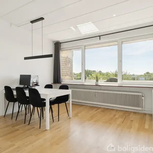 Spisebord med sorte stole står foran et stort vindue, der giver udsigt til grønne træer. På bordet er der en computer. Rummet har lyse trægulve og hvidmalede vægge.