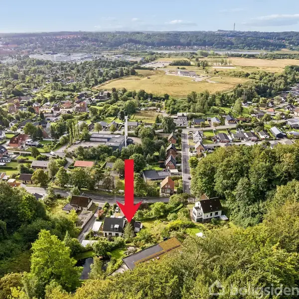 Rød pil peger på et hus omgivet af træer i en kuperet bydel. I baggrunden ses en større byflade med marker og grønt landskab.