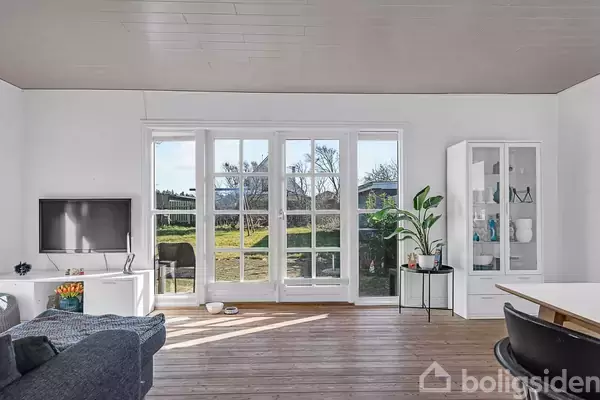 Stue med sofa til venstre, fladskærms-tv og store vinduer mod have. Trægulve og hvid indretning med planter og en reol til højre.